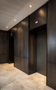 Elevator lobby with architrave and trim complementing the Wenge panelling and travertine floor. - Elevator architrave and panels in V-Grooved PVD coloured stainless steel in Chocolate Vibration. Fenman House, Apartment Building, 9 Handyside St, King’s Cross, London N1C. - Fabrication, V-Grooving and folding of stainless steel by John Desmond Ltd.