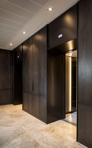 Elevator lobby with architrave and trim complementing the Wenge panelling and travertine floor. - Elevator architrave and panels in V-Grooved PVD coloured stainless steel in Chocolate Vibration. Fenman House, Apartment Building, 9 Handyside St, King’s Cross, London N1C. - Fabrication, V-Grooving and folding of stainless steel by John Desmond Ltd.