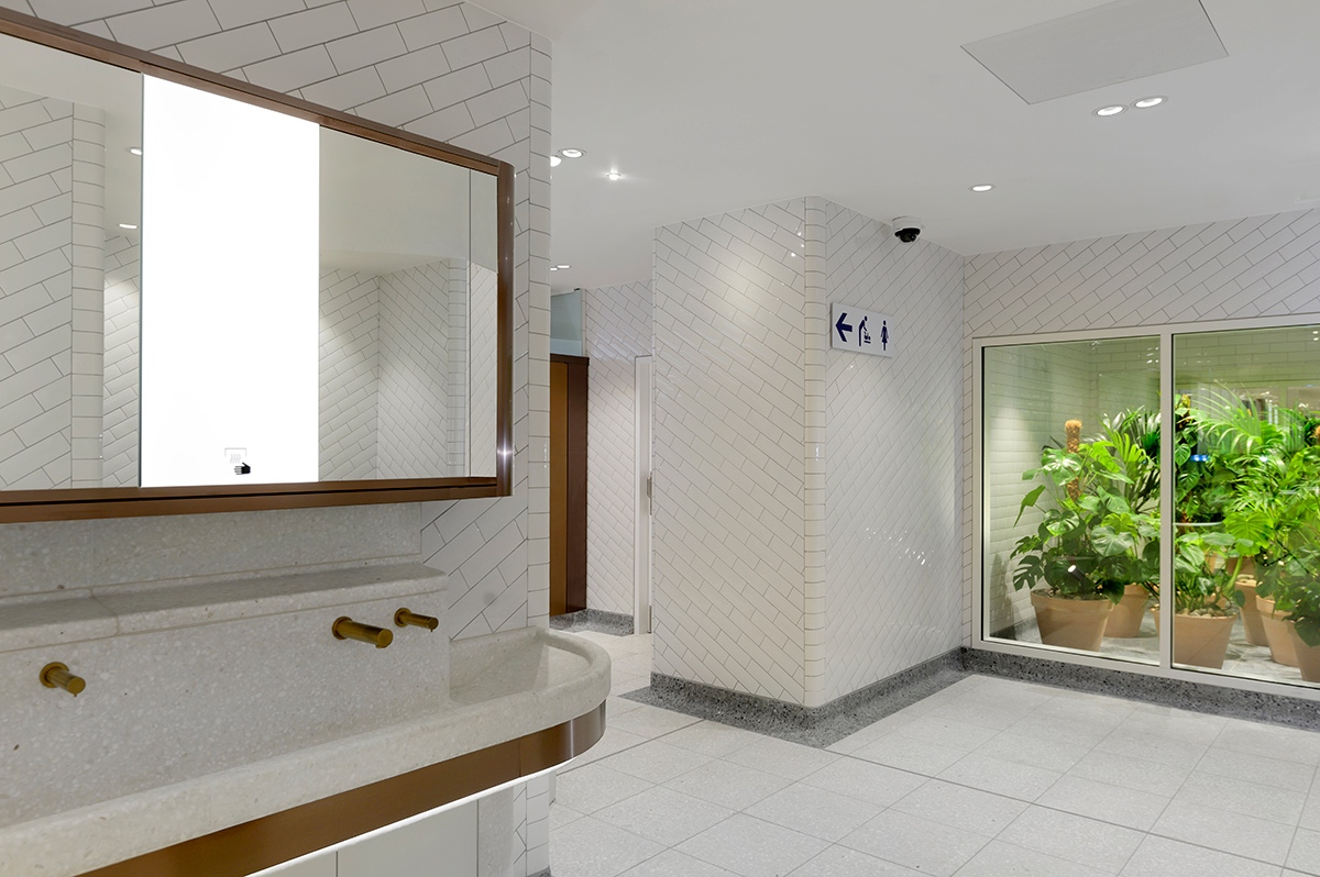 Interior of upgraded WCs Victoria Station, London showing bronze PVD stainless steel taps with matching mirror frame and horizontal banding around communal basin.