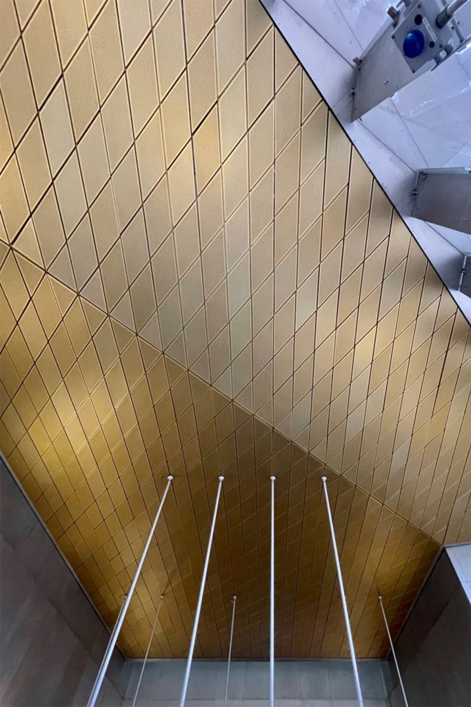 Showing the gold, or brass-coloured, anodised aluminium diagonally-set ceiling panels to the Wood Lane Underground Station, London. Architectural design by Ian Ritchie Architects. Ceiling by John Desmond Ltd.